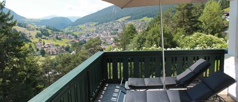 A balcony with two lounge chairs and a large umbrella, overlooking a scenic view of a town and mountains.