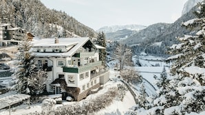 A two-story chalet with a balcony, surrounded by snow-covered trees and mountains.