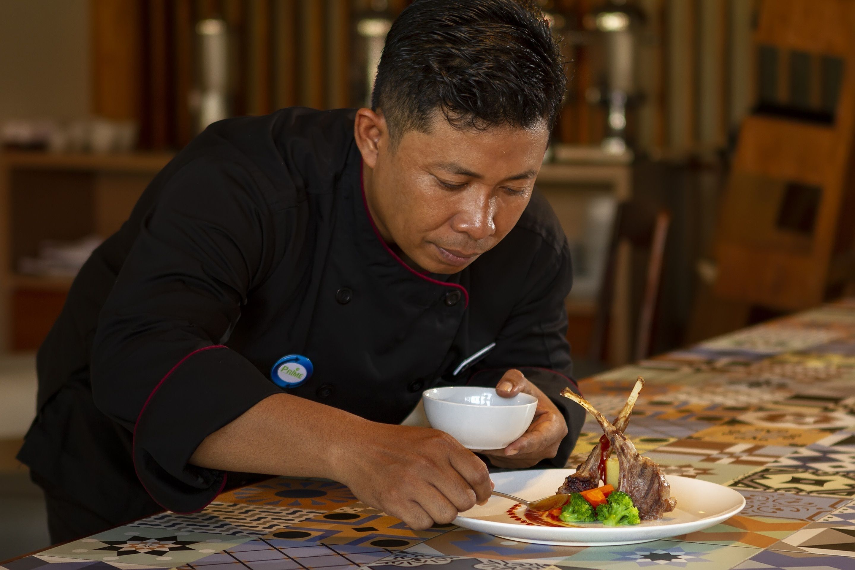 Un chef con uniforme negro y placa sirve un plato con chuletas de cordero, verduras y salsa sobre un mantel estampado.
