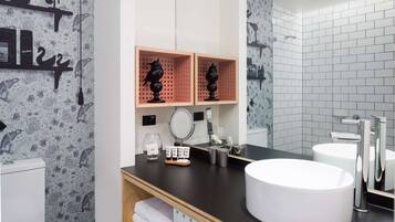Un baño moderno con encimera negra, lavabo blanco y espejo. Hay repisas con artículos de aseo y un perchero.