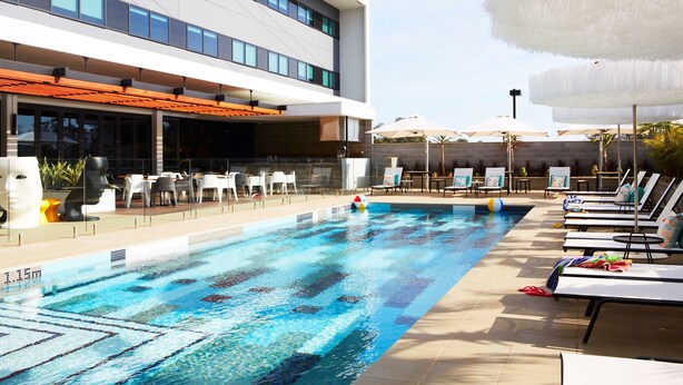 A swimming pool with lounge chairs and umbrellas, a modern building with large windows, and outdoor seating. - Atura Blacktown (Prospect)