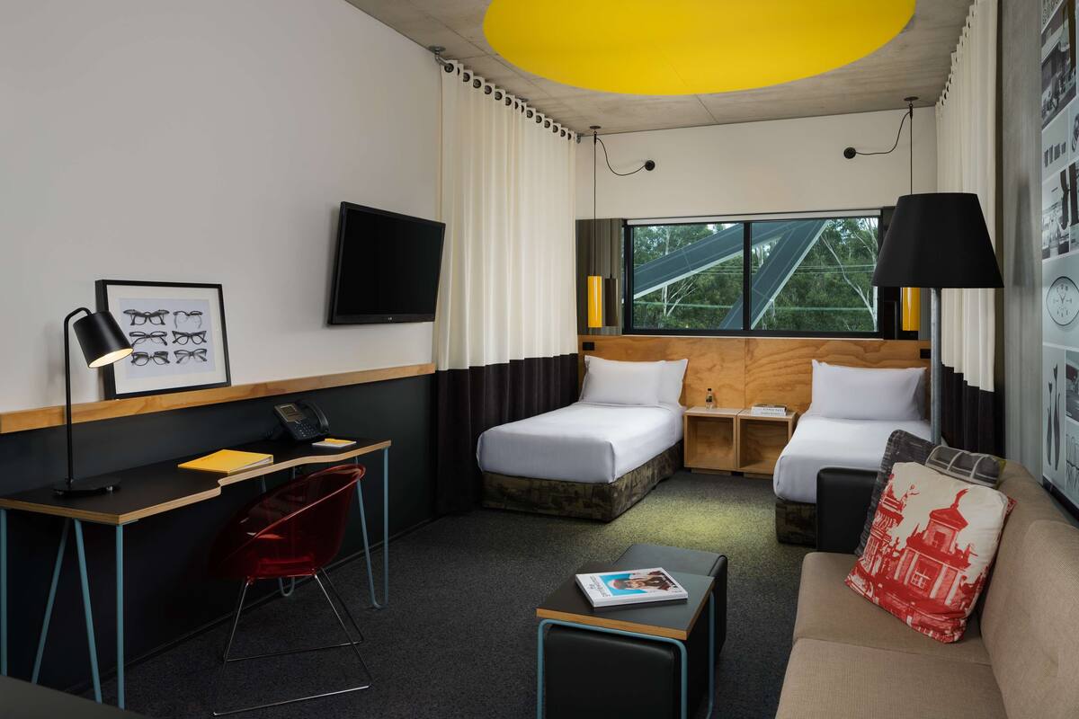 A hotel room with a bed, a desk, a chair, a sofa, a TV, and a window with a view of trees.