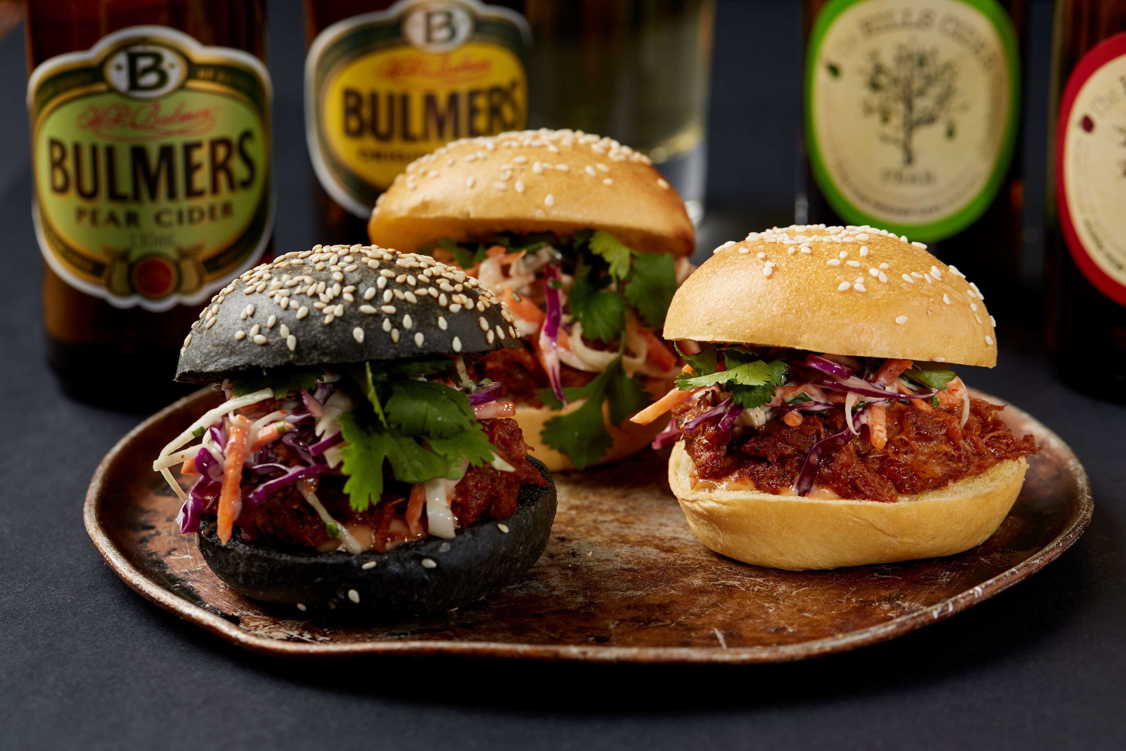 Deux hamburgers sur une assiette en bois, avec une bouteille de cidre à la poire Bulmers en arrière-plan.