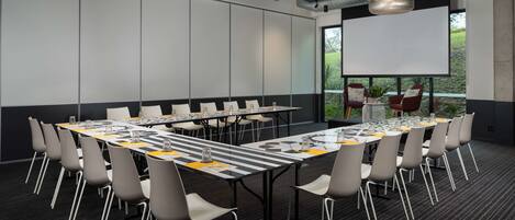 Un salón de conferencias con una gran mesa, sillas, una pantalla de proyector y vistas a zonas verdes al exterior.