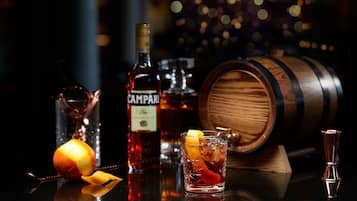 A bar setting with a glass of whiskey, a bottle of Campari, a wooden barrel, and a glass pitcher.