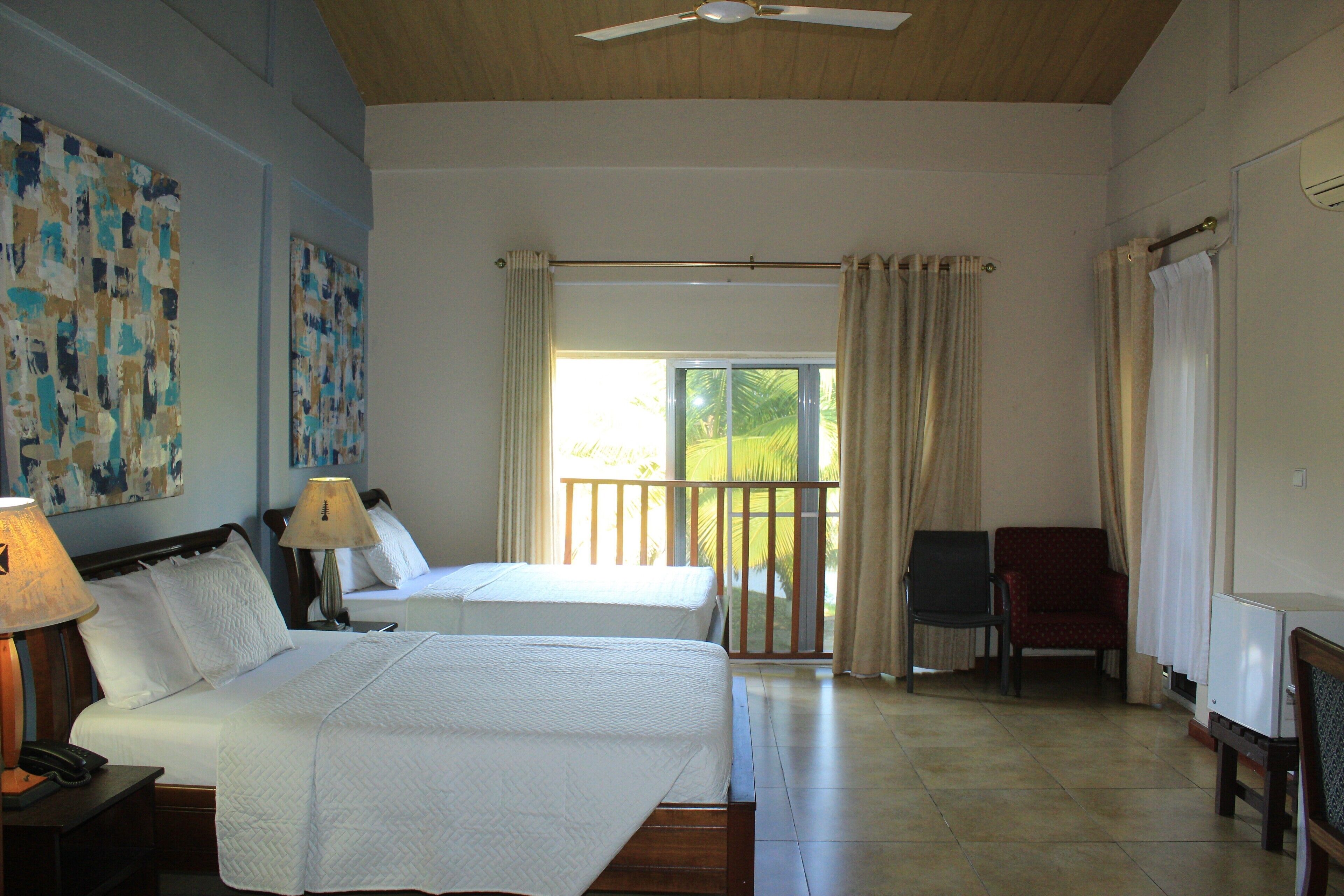 A hotel room with two beds, a balcony with curtains, a chair, and a desk with a lamp.