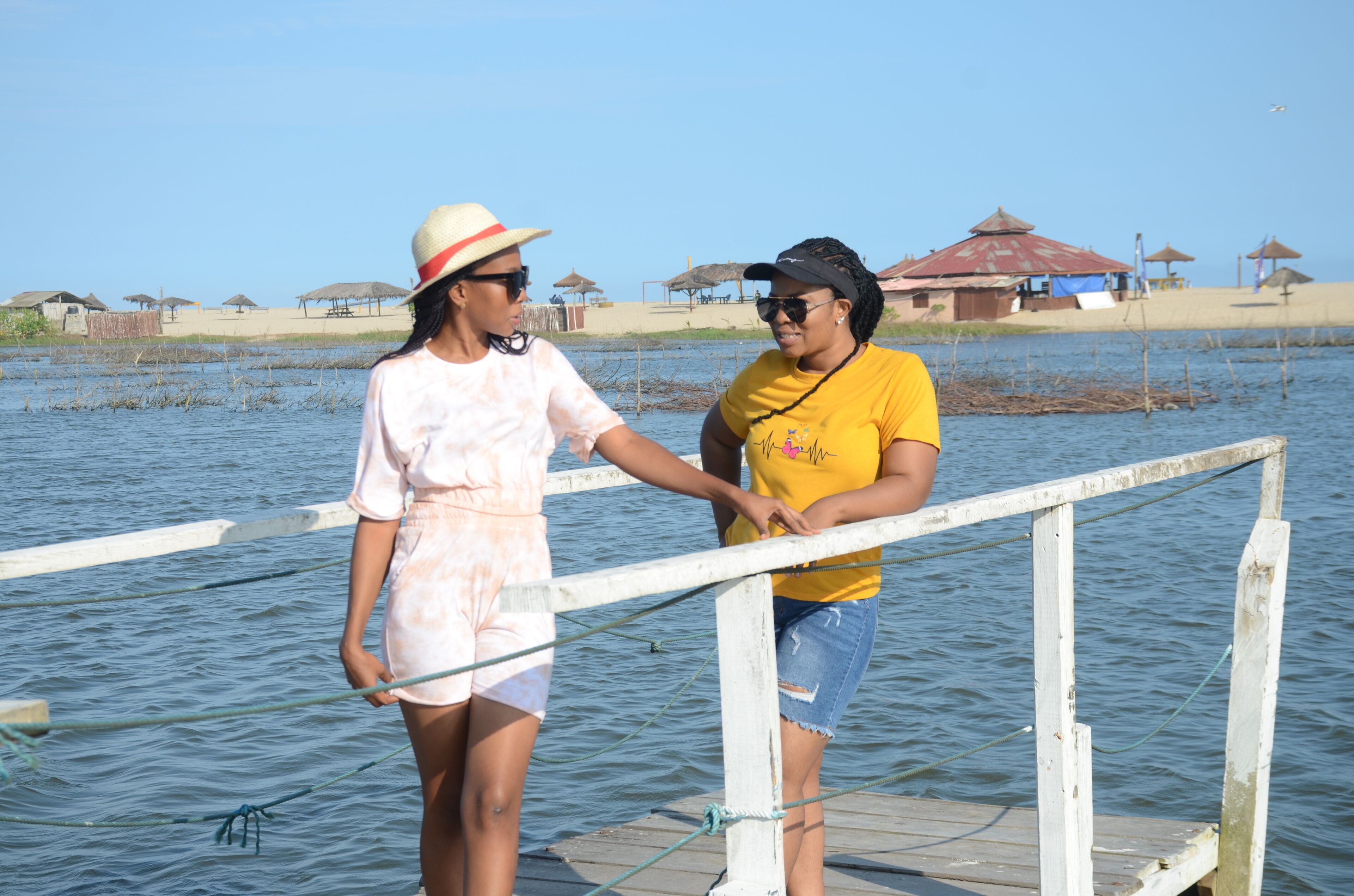 Two people standing on a wooden dock by a body of water with thatched-roof structures in the background.