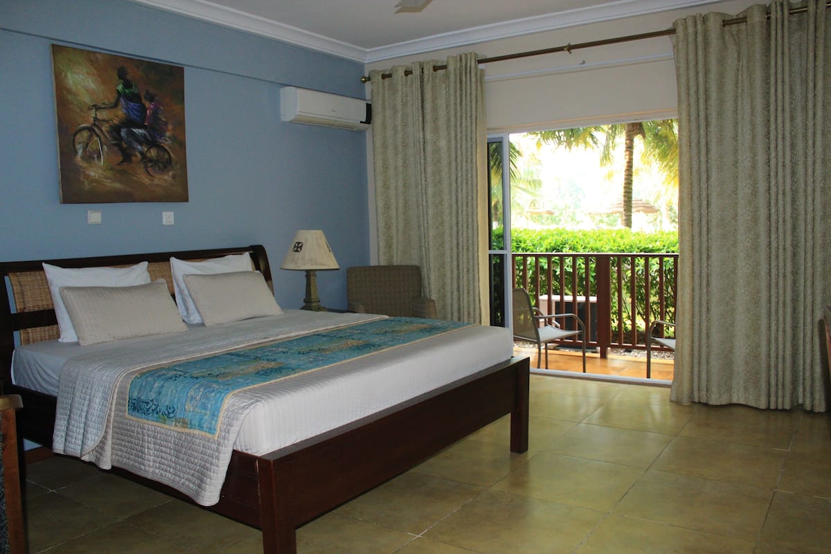 A bedroom with a bed, a painting, an air conditioner, a lamp, and a view of a balcony with a chair.