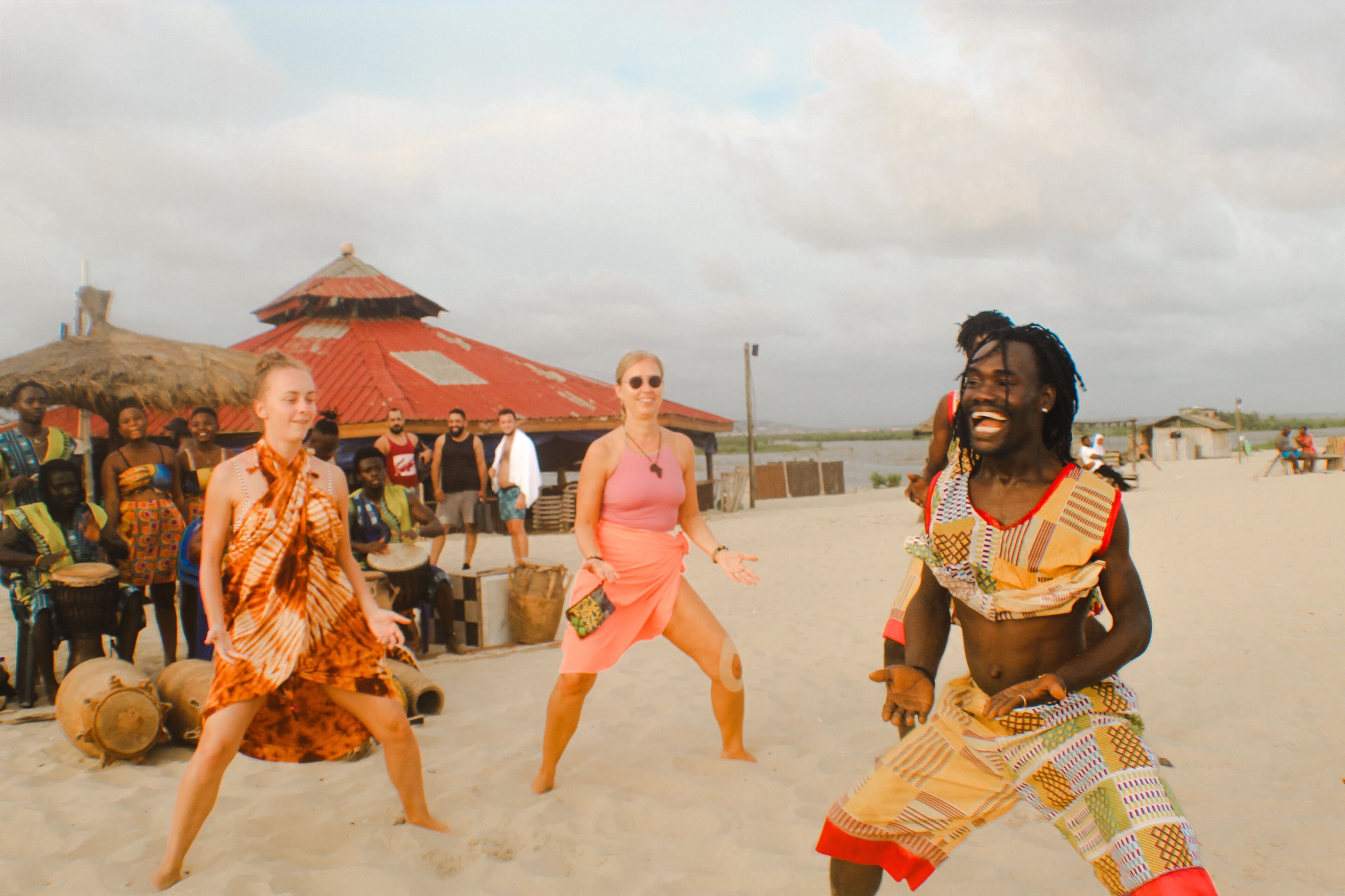 Escena de playa con gente bailando, una cabaña con techo de paja y una playa arenosa.