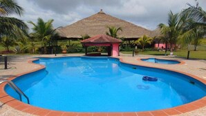 A swimming pool with a tiled deck, surrounded by palm trees and a thatched-roof structure. - Bojo Beach Resort (Bortianor)