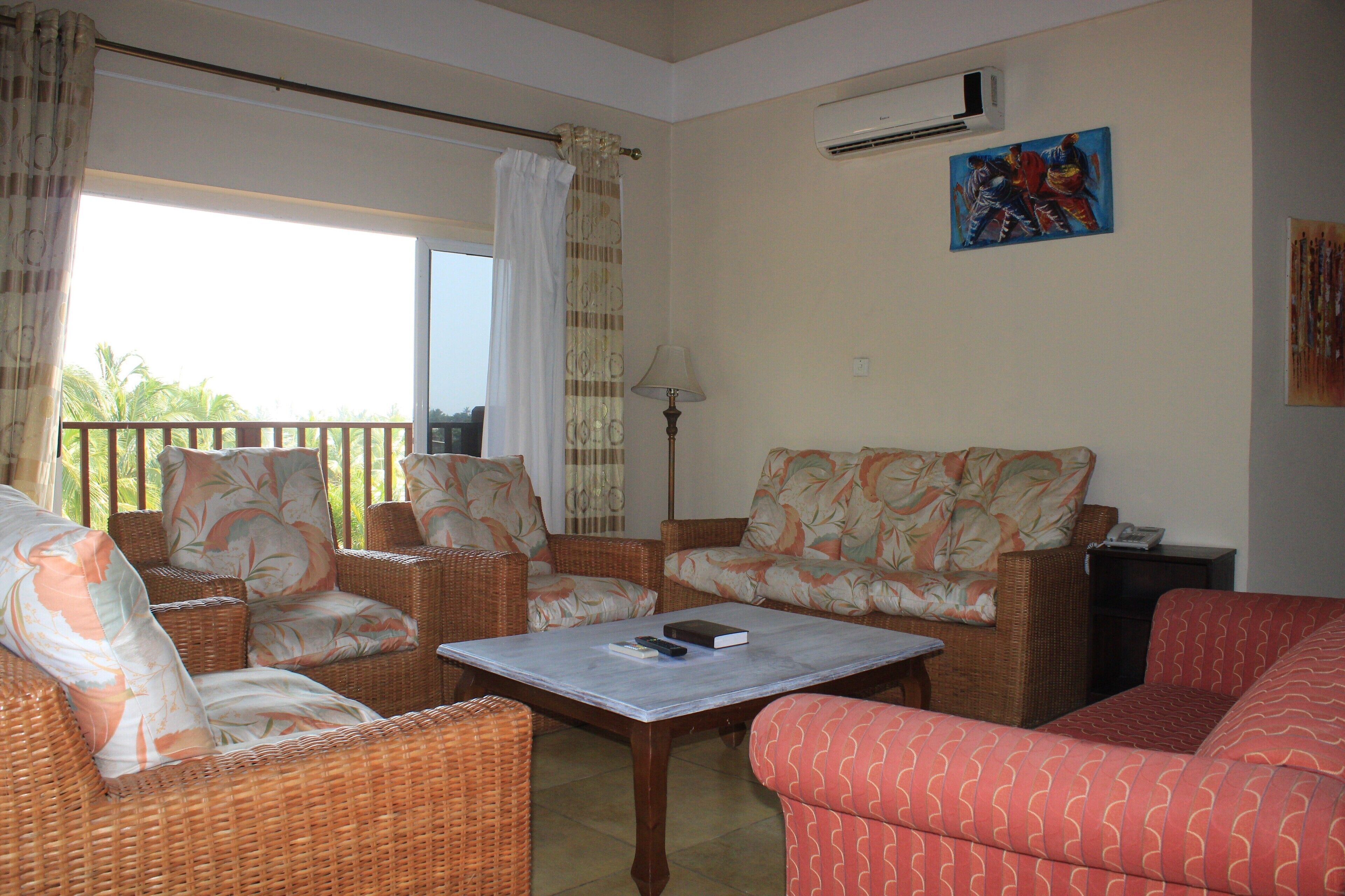 A living room with wicker furniture, a table with a book and remote, and a view through a window.