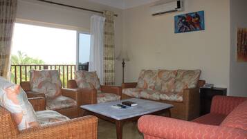 A living room with wicker furniture, a table with a book and remote, and a view through a window.
