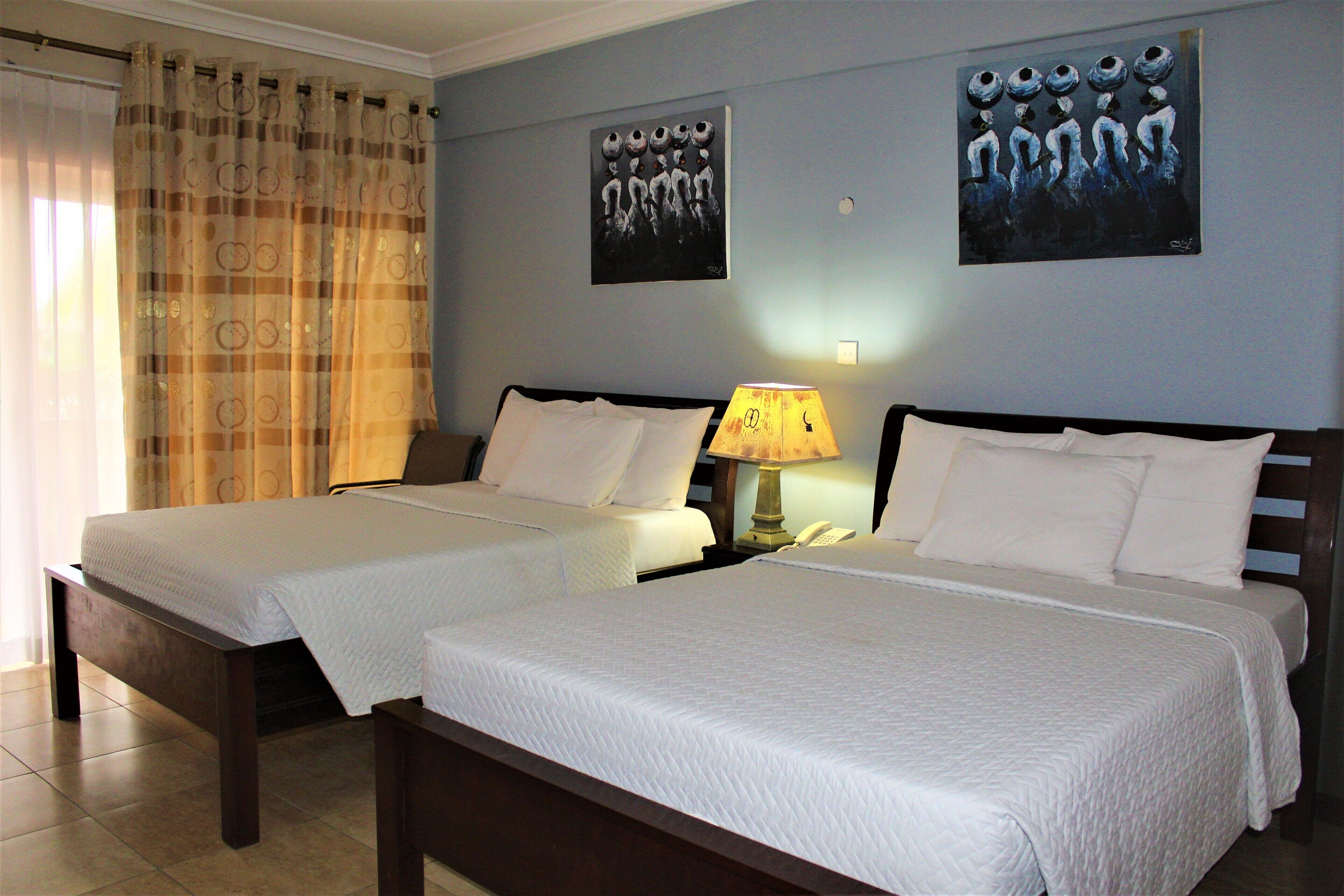 A hotel room with two single beds, a nightstand with a lamp, and two wall-mounted paintings.