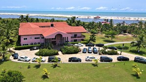 A pink building with a red roof, surrounded by a parking lot and palm trees, overlooking a beach. - Bojo Beach Resort (Bortianor)