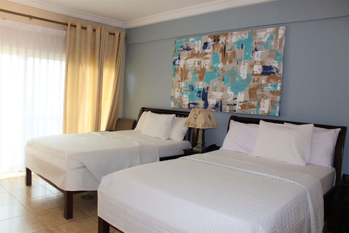 A hotel room with two beds, beige curtains, a wall painting, and a bedside lamp.