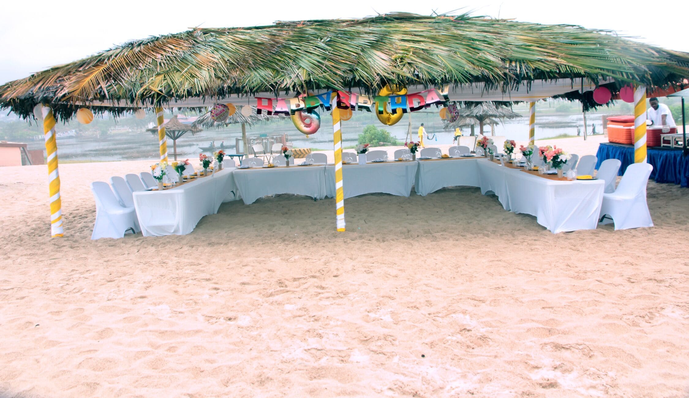 Montaje para un evento en la playa, con techo de paja, mesas y sillas blancas, decorado con globos y flores.