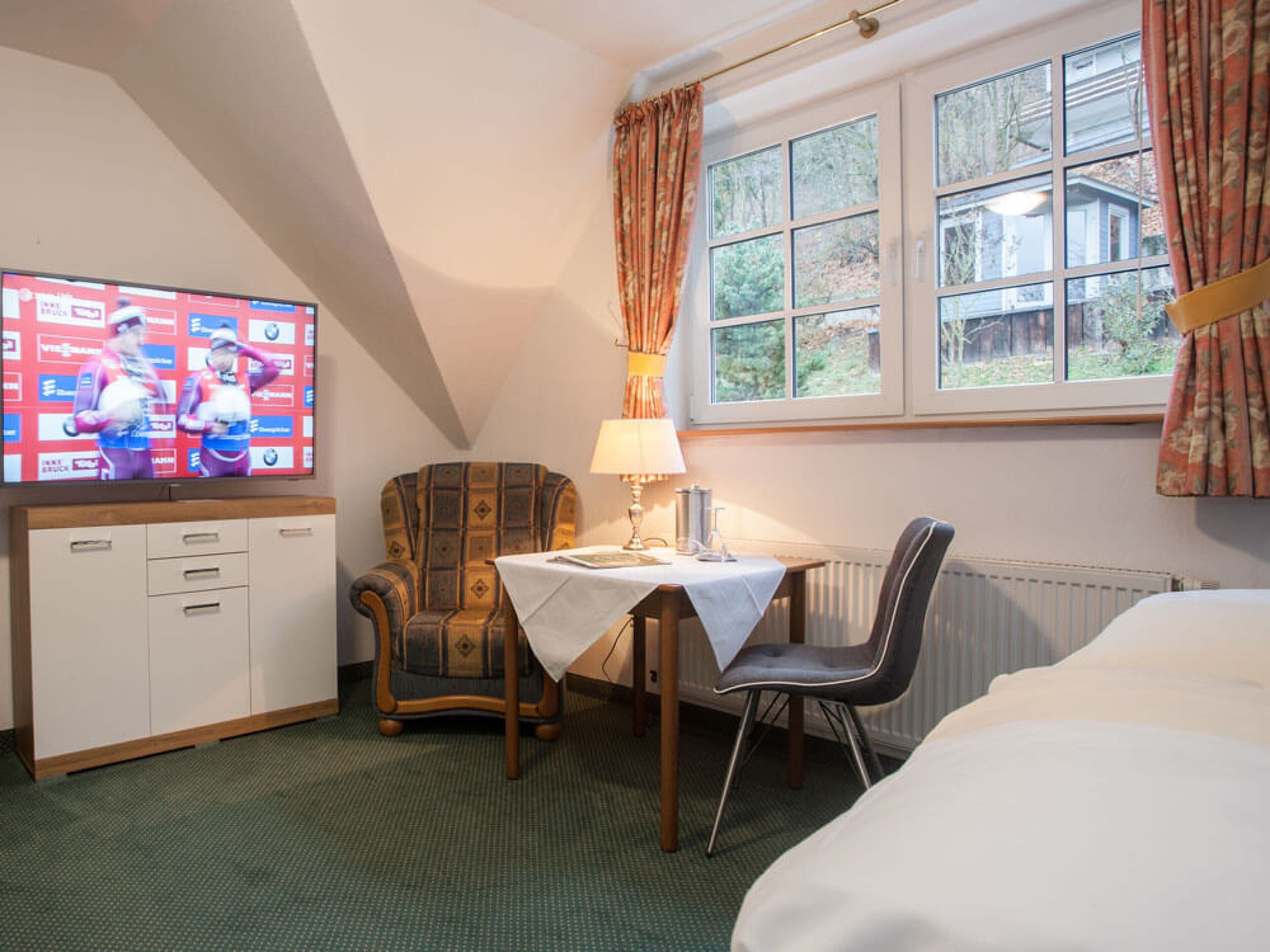 Une chambre d’hôtel équipée d’un téléviseur diffusant un événement sportif, d’une table à manger avec une lampe, d’un fauteuil à motifs, d’une chaise moderne et d’un lit.