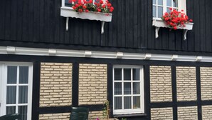 A building with black exterior walls, white window frames, and a brick pattern. There are two windows with flower boxes containing red flowers. A green chair and a table are visible in front of the building. - Landhotel Menke (Brilon)