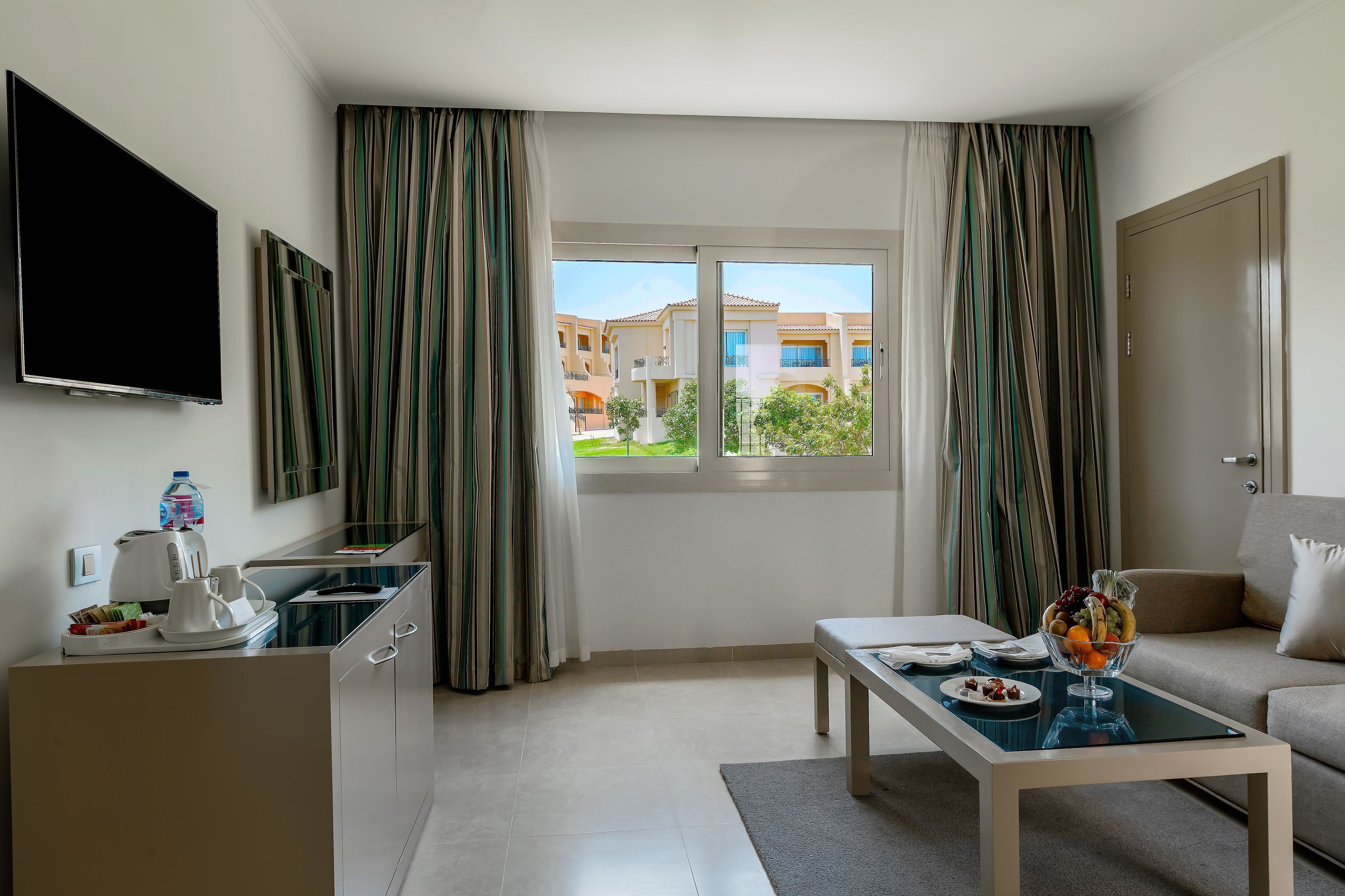 A modern hotel room with a kitchen area, a dining table with a fruit bowl, and a sofa.