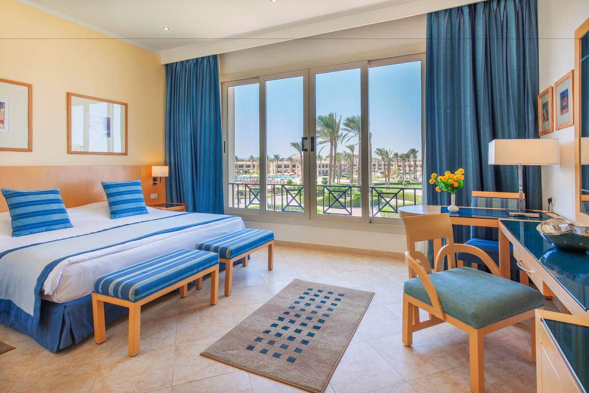 Een hotelkamer met een groot bed, een bureau, een stoel en uitzicht op een zwembad en palmbomen.