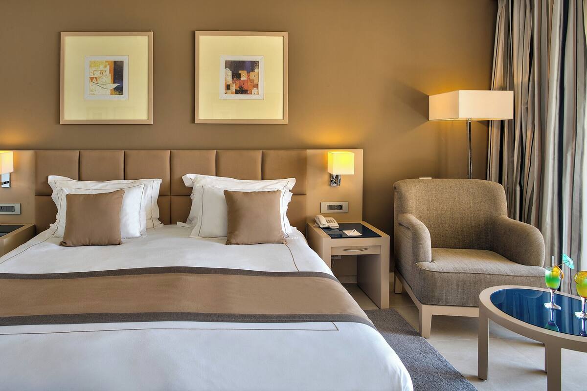 Een hotelkamer met een groot bed, twee bedlampen, een stoel, een klein tafeltje met drankjes en twee ingelijste foto's aan de muur.