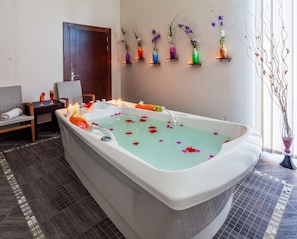 Ein Wellnessraum mit einer großen Badewanne, gefüllt mit Wasser und darin schwimmenden Blumen, einem kleinen Tisch mit Getränken, einem Sessel und einer Wand mit Regalen, auf denen farbenfrohe Vasen stehen.