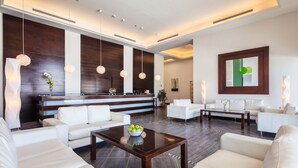 Un hall d’entrée moderne avec un comptoir de réception, des canapés blancs, une table basse sur laquelle est posé un saladier rempli de fruits, des luminaires suspendus et un grand tableau abstrait.