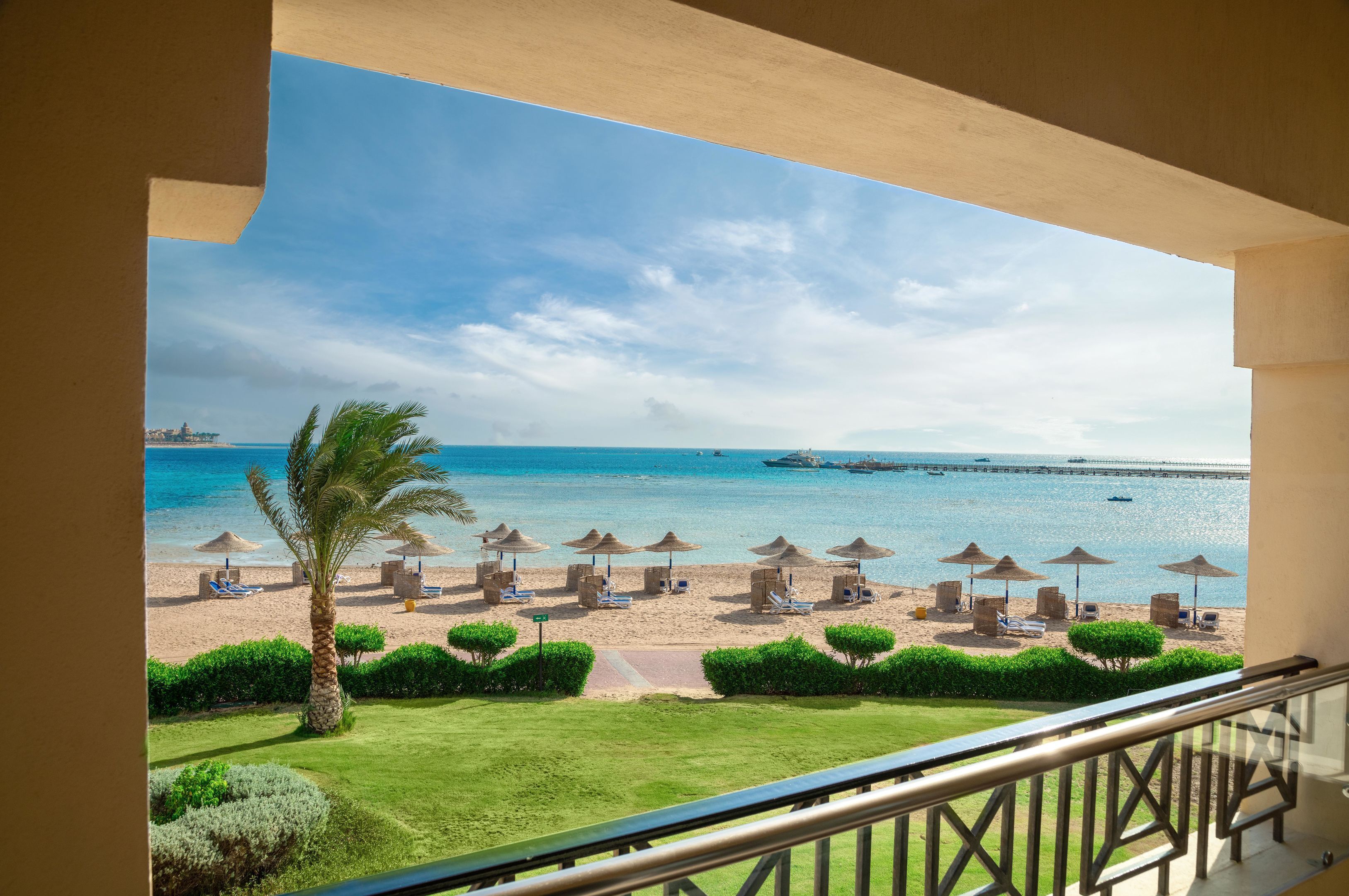 A balcony with a view of a beach, sun loungers, umbrellas, and a palm tree.