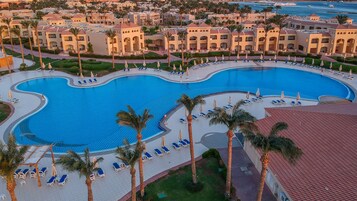 A large, curved swimming pool surrounded by palm trees and lounge chairs, with a view of hotel buildings in the background.