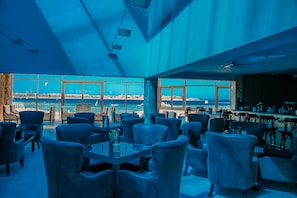 A spacious indoor area with a large window overlooking a coastal view, featuring multiple seating arrangements and a bar area. - Cleopatra Luxury Resort Makadi Bay (Makadi Bay)