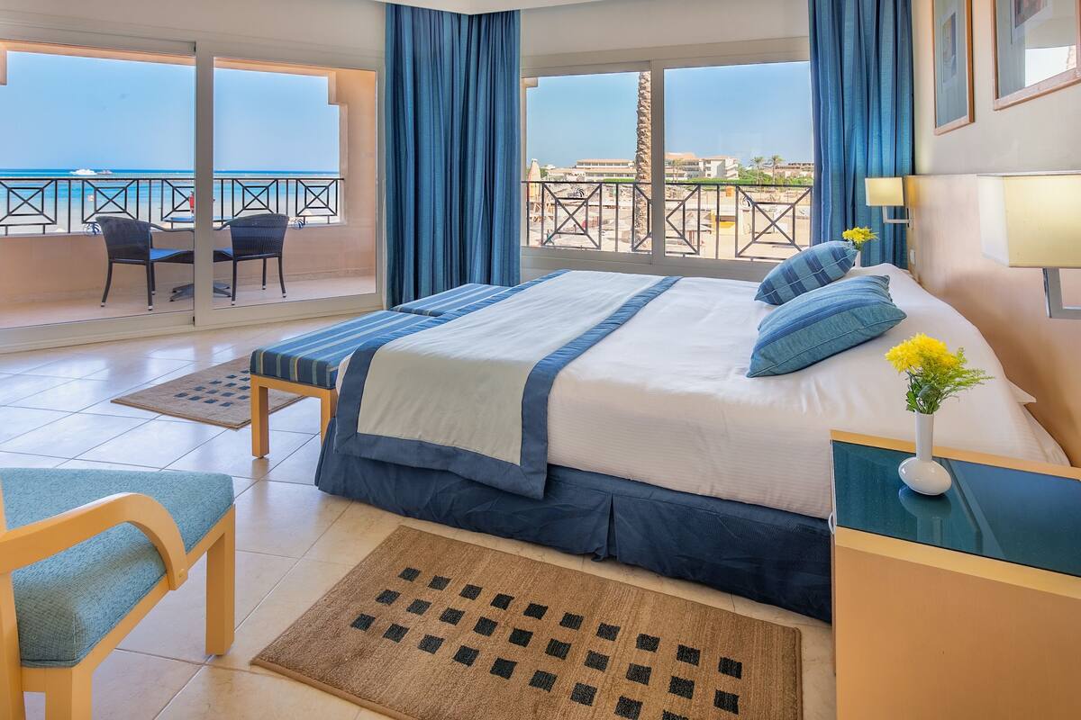 Een hotelkamer met een groot bed, een stoel, een tafeltje, een nachtkastje, een raam met uitzicht op zee, en een balkon met een stoel en tafel.