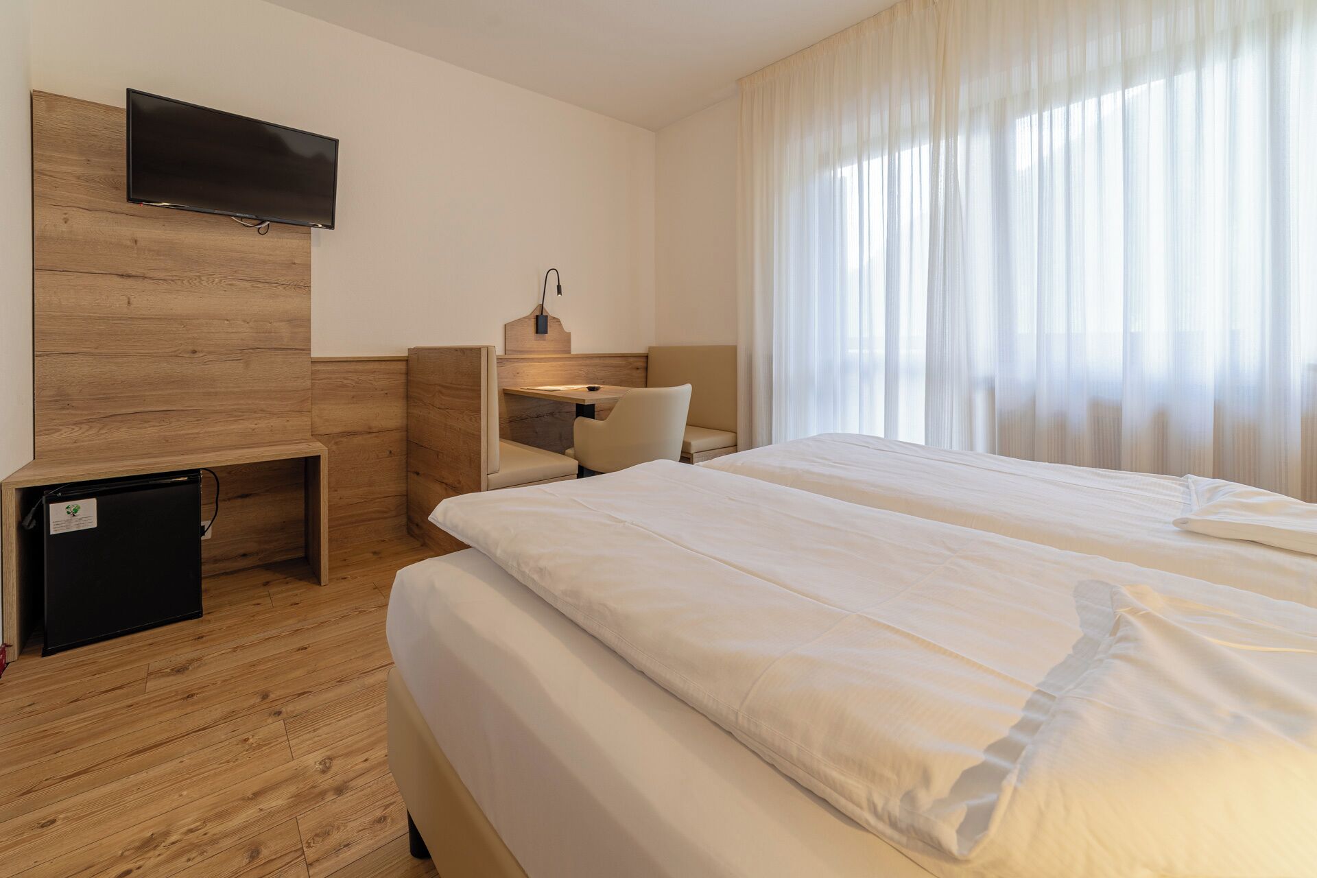 A hotel room with a bed, a desk, a chair, a TV mounted on a wooden panel, and a small fridge.