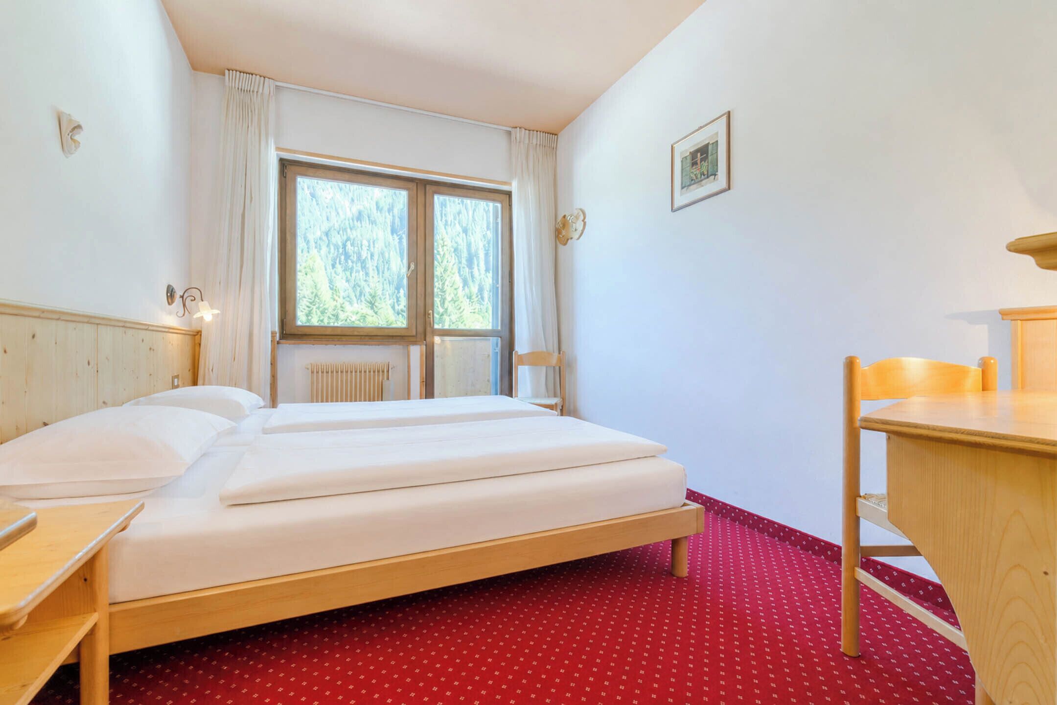 A hotel room with a wooden bed, a desk, a chair, a window with curtains, and a radiator.