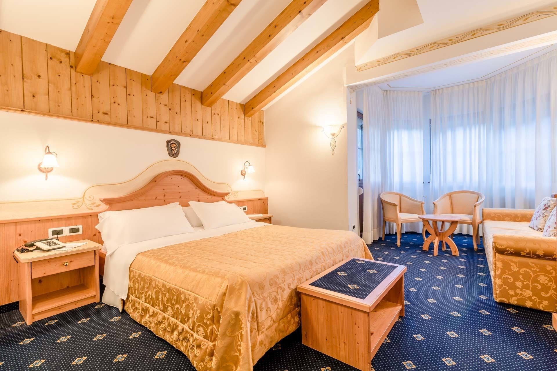 Une chambre d’hôtel avec un lit doté d’une tête de lit en bois, deux tables de chevet avec des lampes, une petite table d’appoint en bois, une moquette bleue à motifs et un coin salon avec deux fauteuils et une petite table.