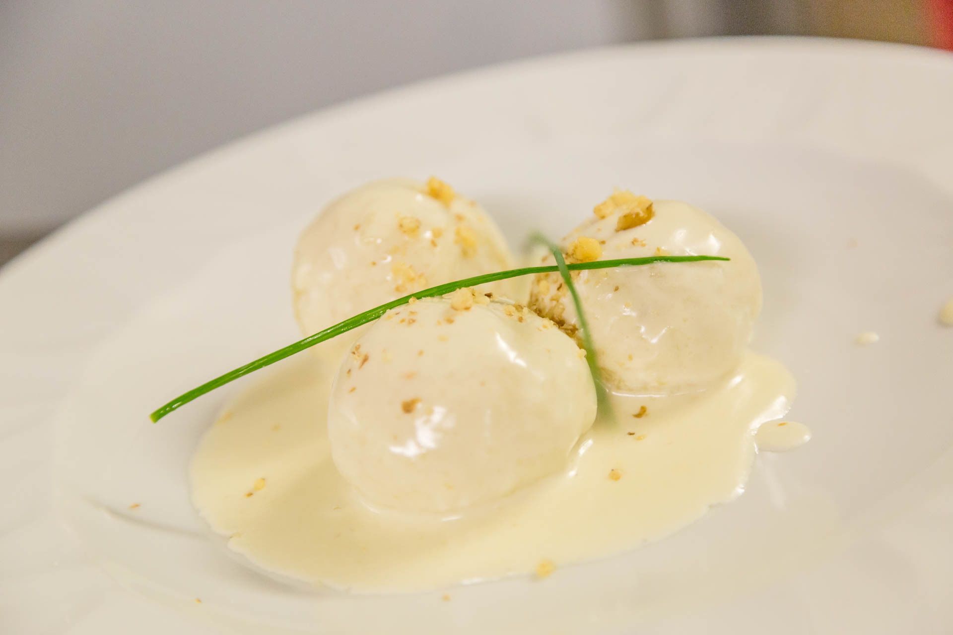 A plate with three round, white dumplings in a creamy sauce, garnished with a green chive.
