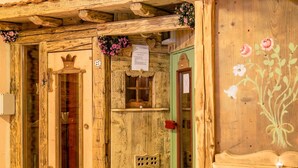 A rustic wooden building with a green door, a window with a decorative wooden frame, and a small shelf with a flower arrangement. - Pineta Pastry Hotel (Rocca Pietore)