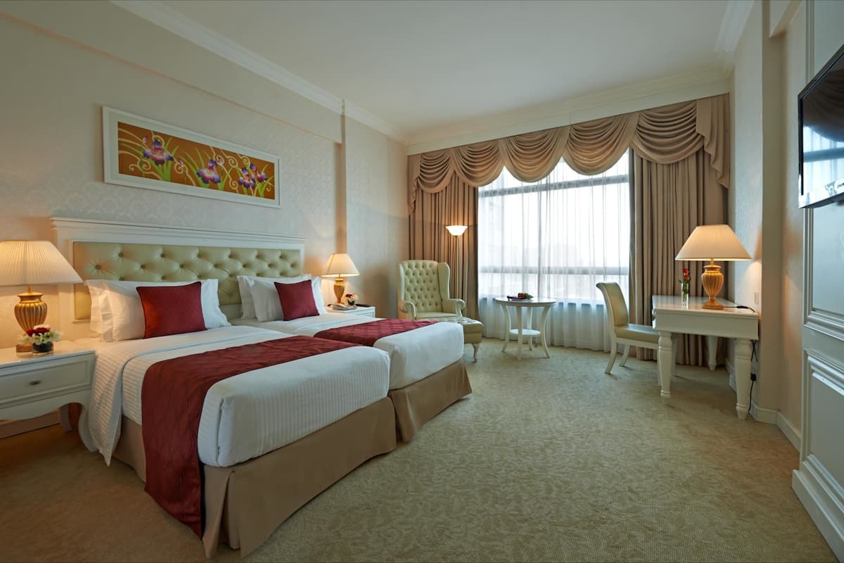 A hotel room with two beds, a sitting area with a chair and table, and a large window with curtains.