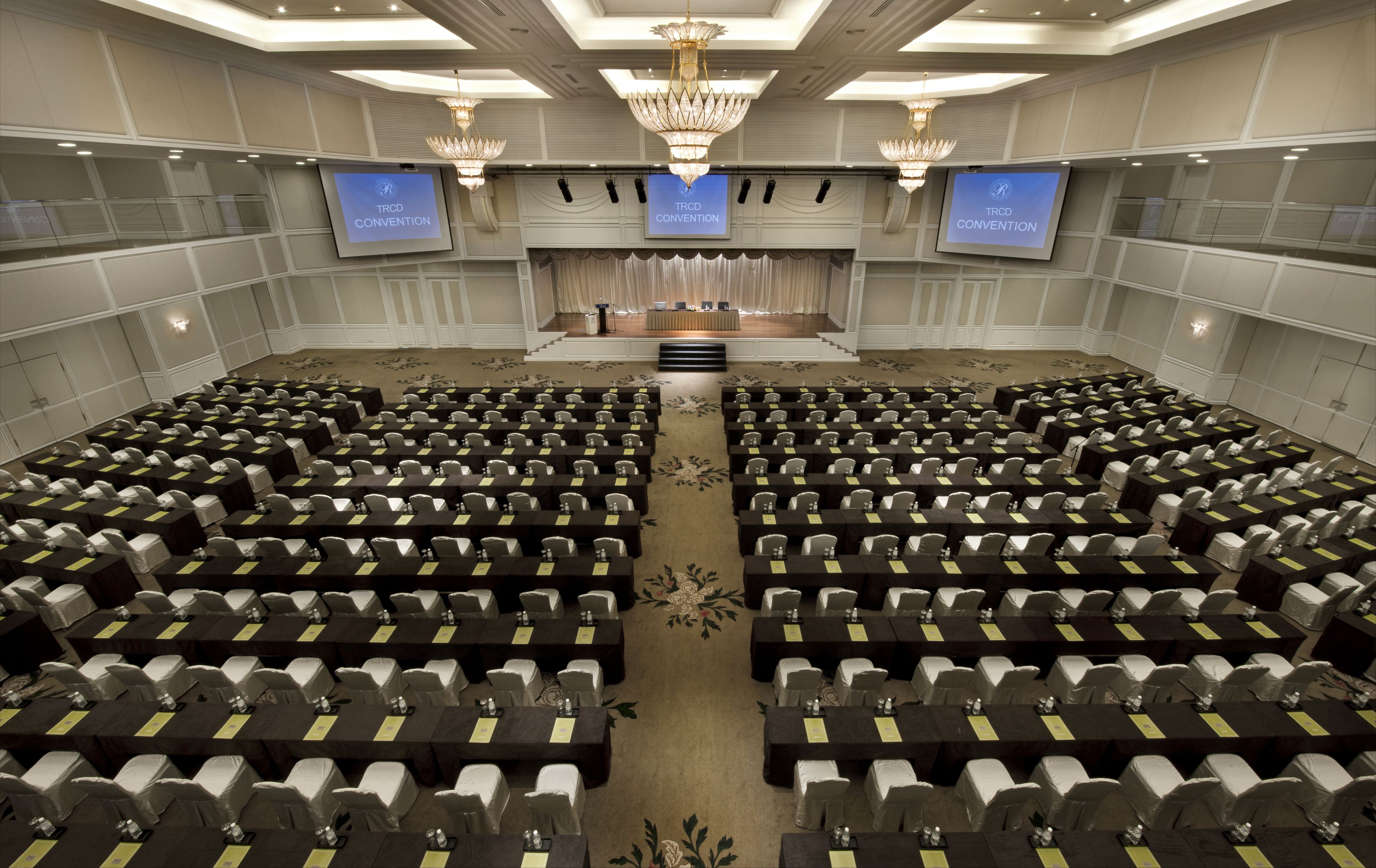 A conference room with rows of tables and chairs, a stage with a podium, and three screens displaying 'TRCD CONVENTION'.