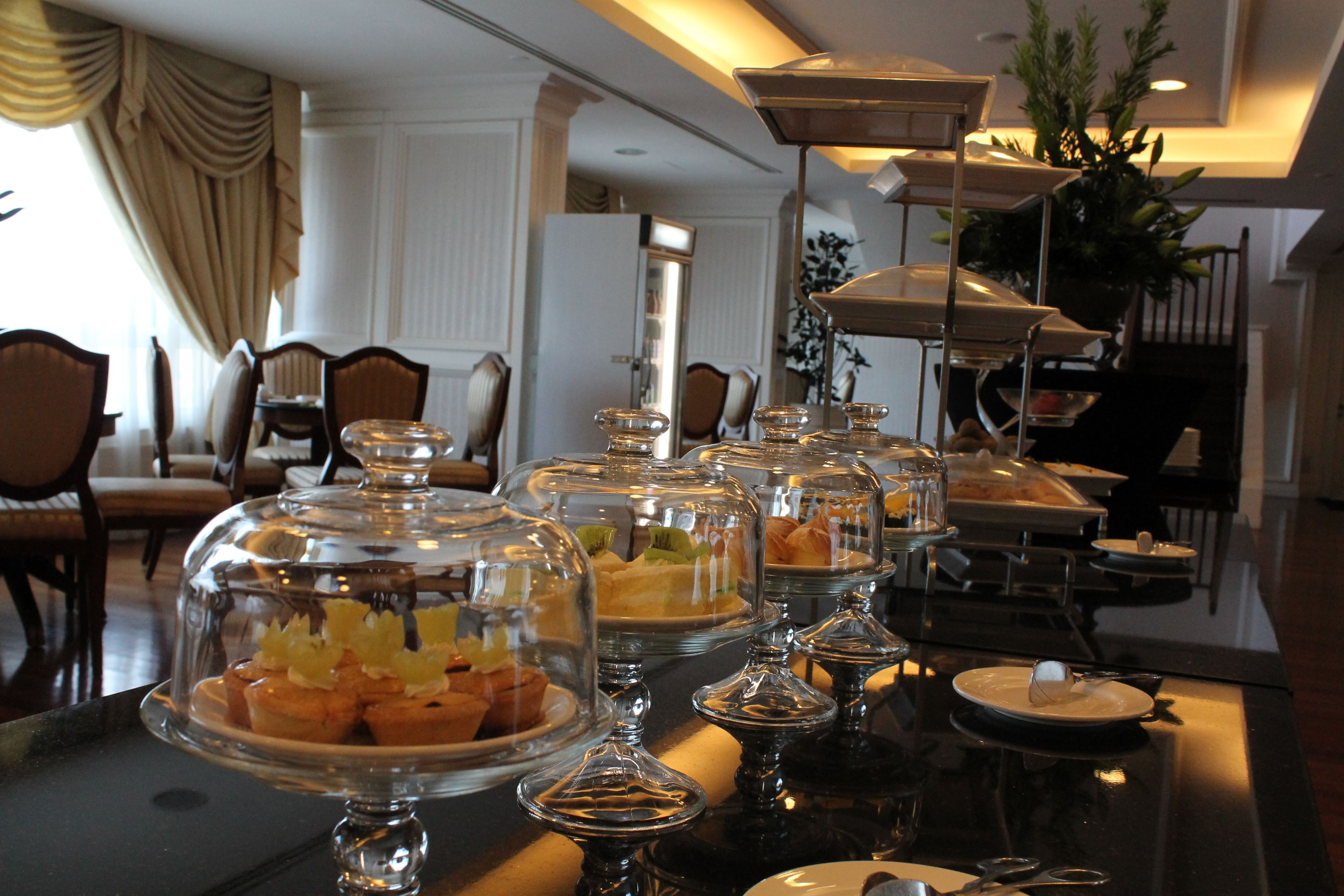 A buffet setup with various food items under glass domes, a dining area with chairs and tables, and a plant in the background.