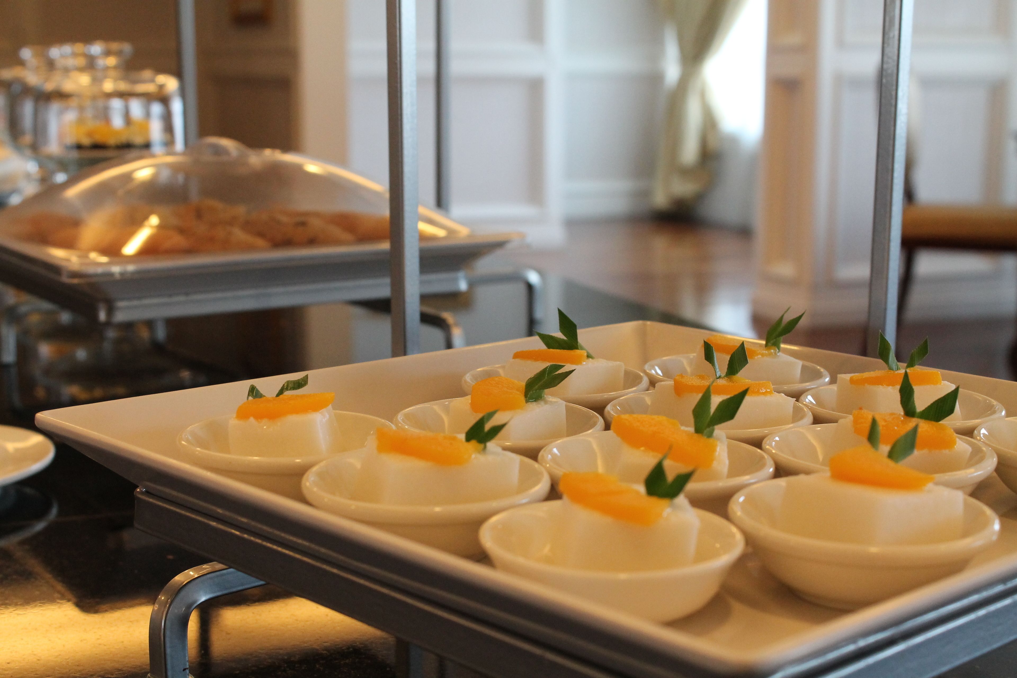 A buffet setup with white bowls containing food garnished with orange slices and green leaves, arranged on a metal serving cart.