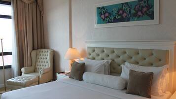 A hotel room with a large bed, a tufted headboard, a reclining chair, a bedside table with a lamp, and a floral painting on the wall.