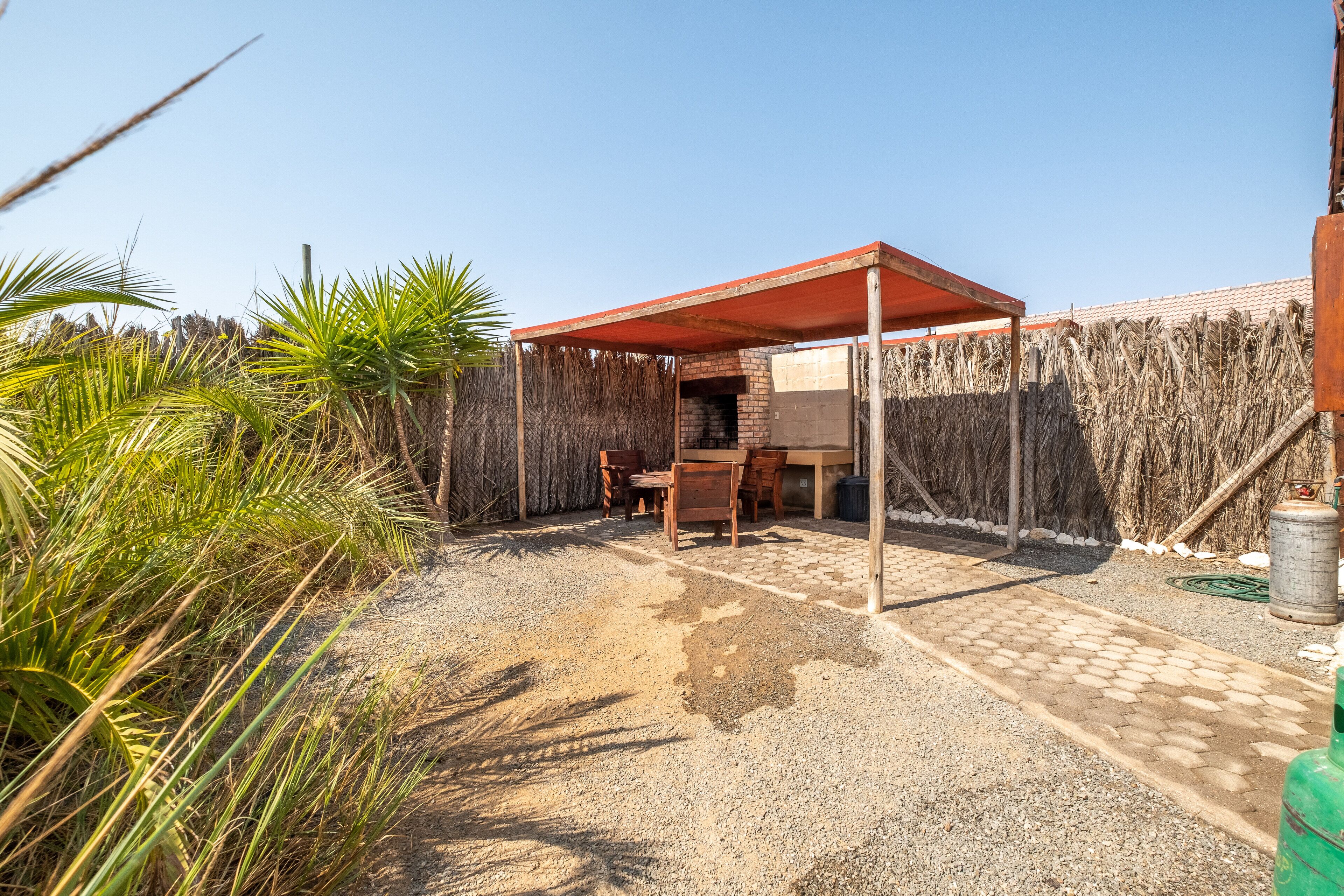 A covered outdoor seating area with a table and chairs, surrounded by a wooden fence and palm plants.