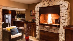 A stone fireplace with a fire, a kitchen area with a refrigerator, and a seating area with a sofa and a vase of flowers. - Gecko Ridge Guest House (Swakopmund)