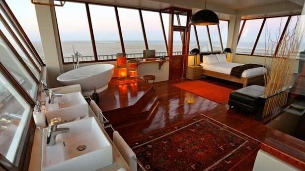 A luxurious cabin with a large window, a bathtub, a desk, and a bed. - Pelican Point Lodge (Walvis Bay)