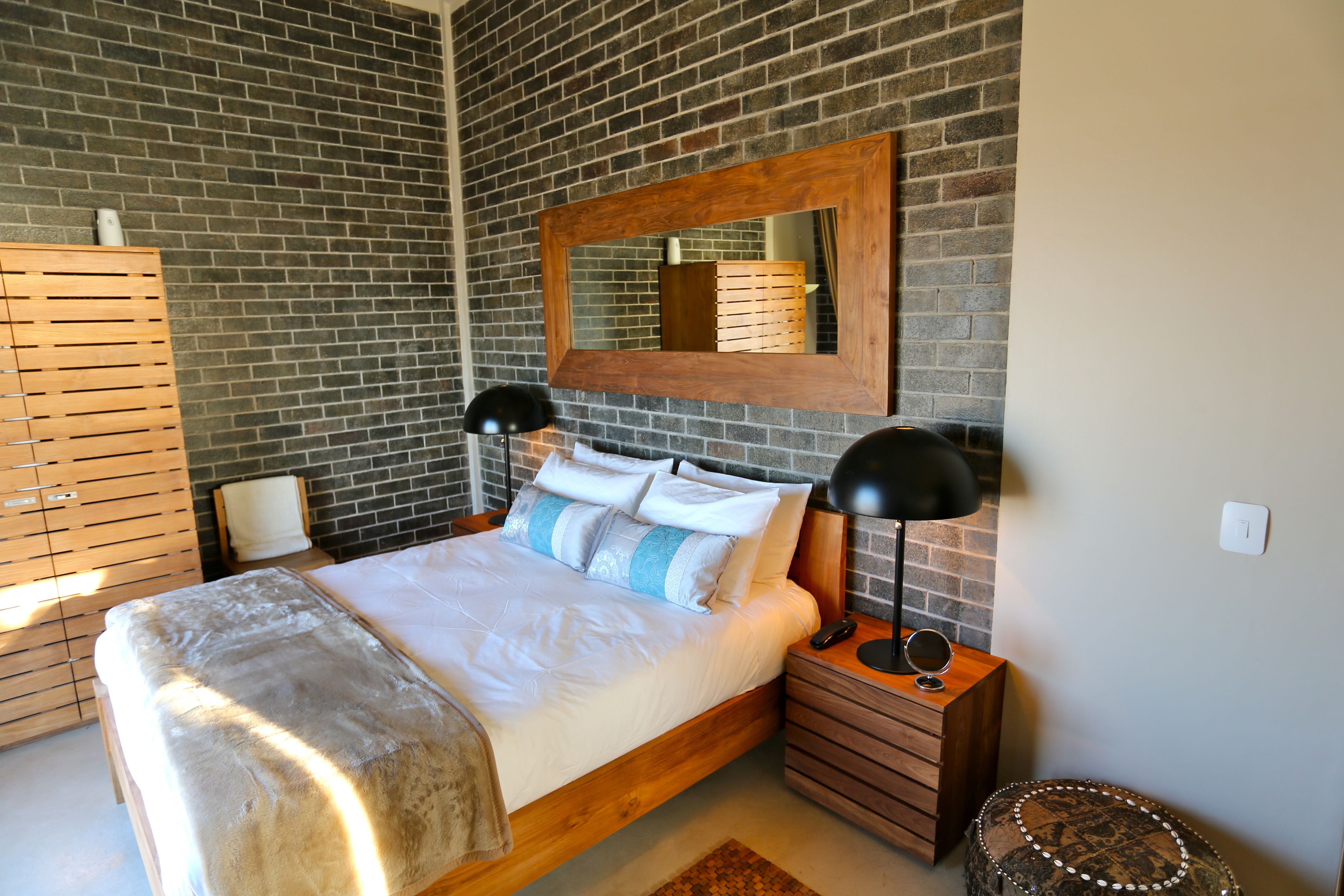 A modern bedroom with a brick wall, a wooden bed frame, a large mirror, bedside lamps, and a small round ottoman.