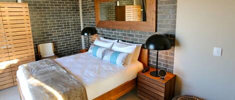 A modern bedroom with a brick wall, a wooden bed frame, a large mirror, bedside lamps, and a small round ottoman.