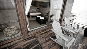 A modern room with a sliding glass door leading to a balcony. The room has a bed, a sofa, and a small table. The balcony has two Adirondack chairs and a table. - Pelican Point Lodge (Walvis Bay)