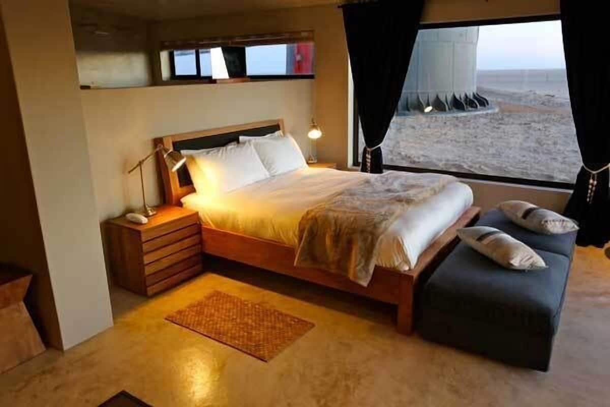 A bedroom with a bed, bedside tables, a bench, a window with a view of the beach, and a shelf with books.
