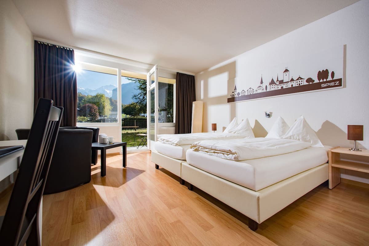 A hotel room with a large bed, a desk, and a chair. There is a window with curtains, a lamp, and a wall decoration.