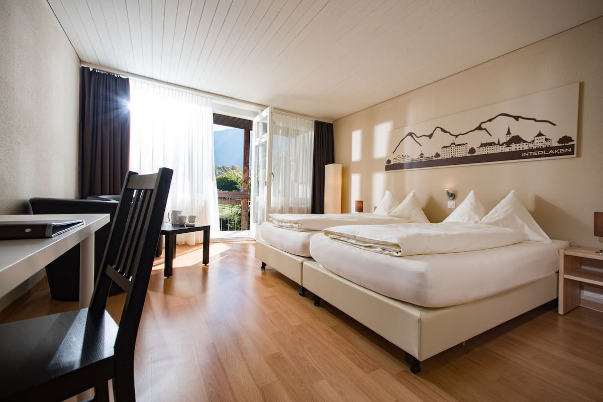 A hotel room with a wooden floor, a desk with a chair, a bed with pillows, a lamp, and a painting of a mountain landscape.